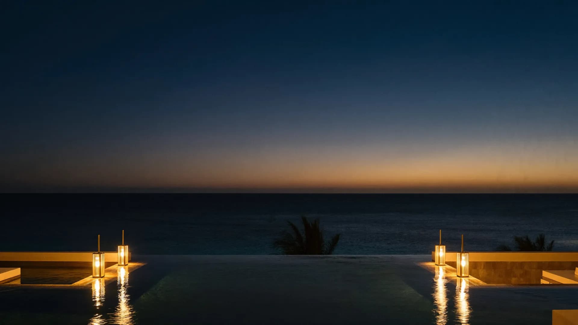 Alberca infinita de un resort de gran lujo fundiéndose con el mar Caribe al anochecer, con lámparas de latón encendidas y palmas contra un cielo azul marino.